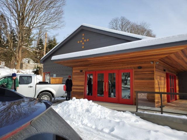 front of the new church with red doors on a wooden and metal panel background. a small wooden cross is in the gable end and snow shows that the move in day is in winter